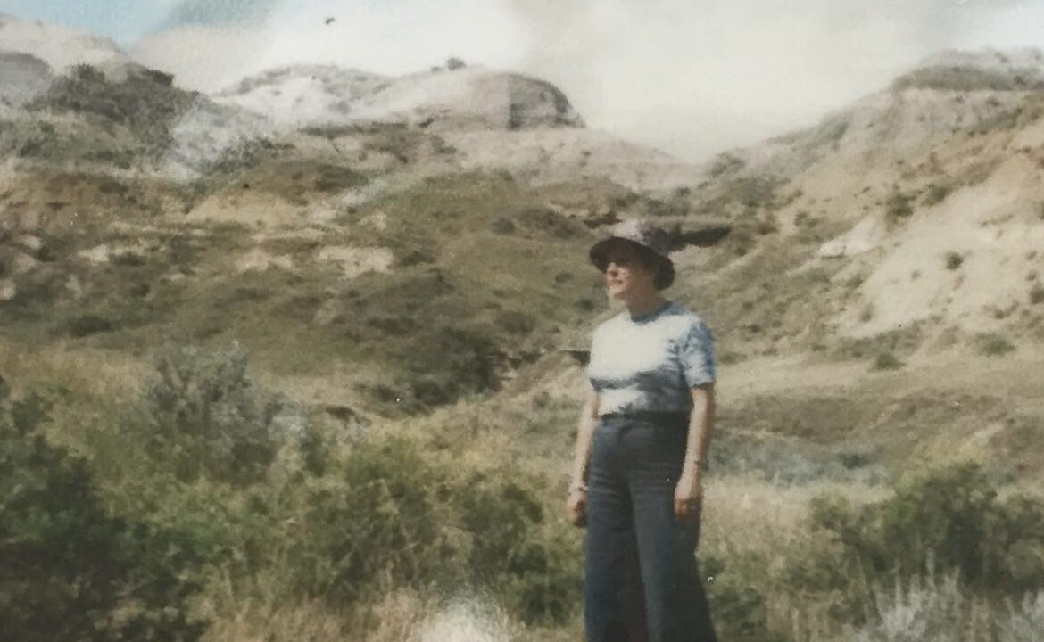 an old photo of a woman wearing a hat standing before arid grass and hills