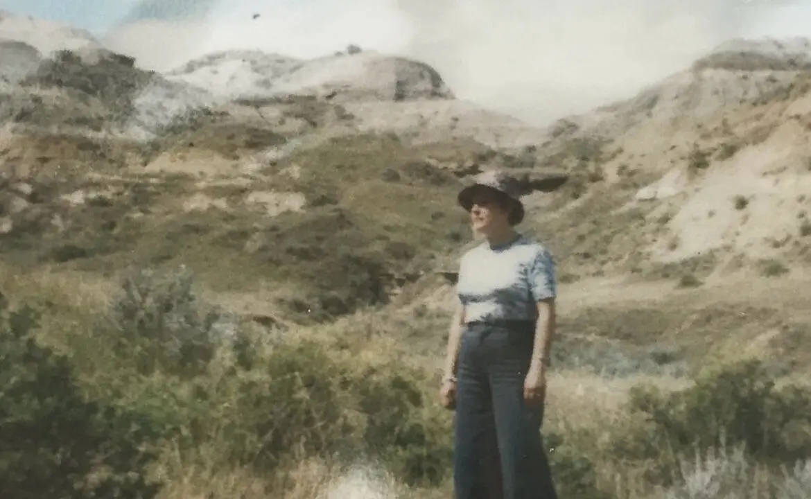 an old photo of a woman wearing a hat standing before arid grass and hills